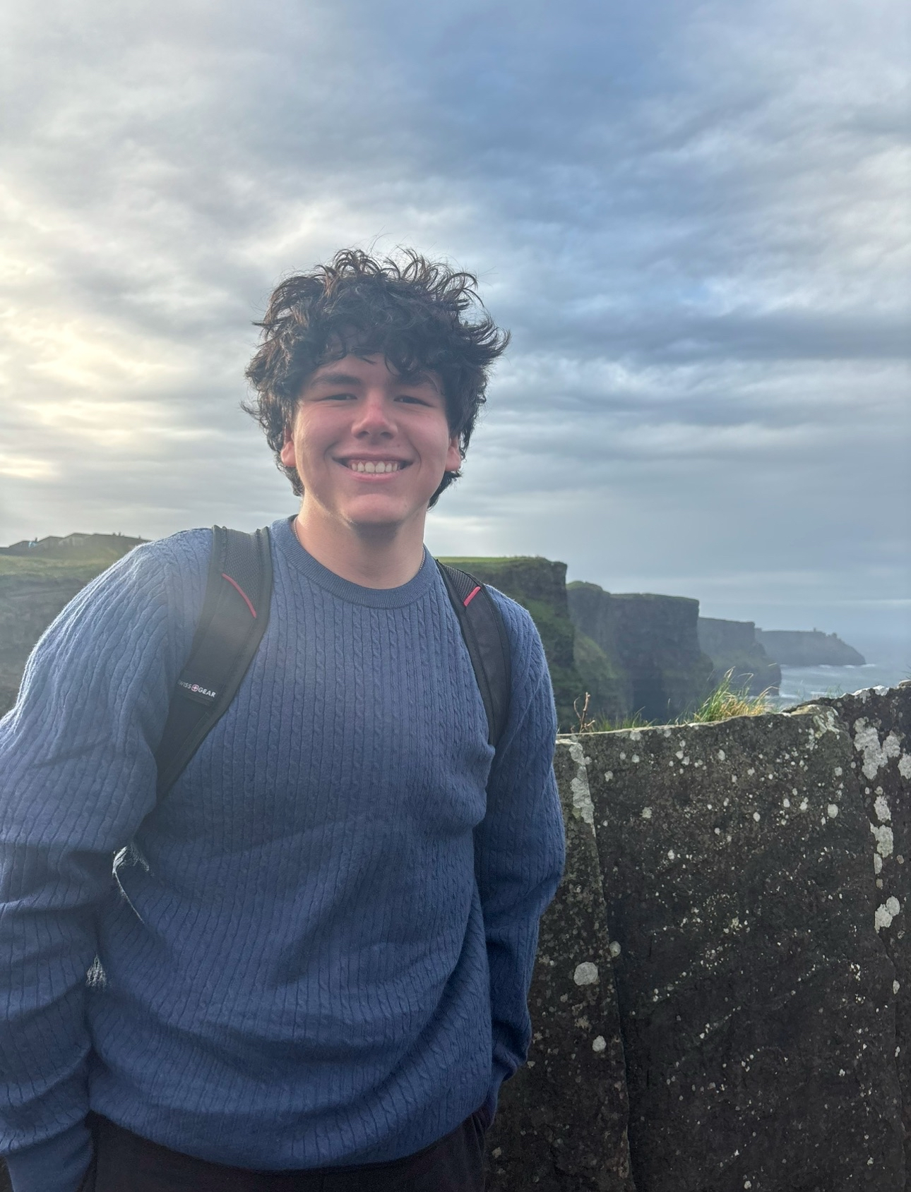 An image of a man with dark hair and a blue sweater, with cliffs and water in the background.